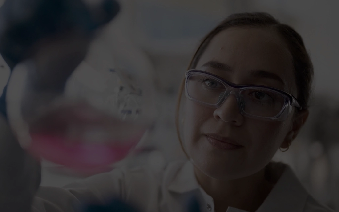 A young woman in a white lab coat looks at a round-bottom flask that is filled with a pink liquid (Photo)