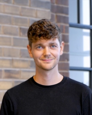 Profile photo of a young man with modern, curly hair in front of a red brick wall and window